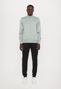 Jachetă verde deschis cu fermoar, guler înalt, manșete elastice și logo. Asortată cu pantaloni negri conici și adidași albi. Textură netedă.