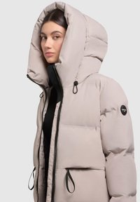 Beige színű puffertél, nagy kapucnival, cipzáros záródással és logóval. Steppelt textúrájában állítható zsinórok találhatók a kapucnin.