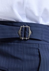 Boucle argentée fixant une ceinture bleu marine à fines rayures sur un pantalon assorti, portée avec une chemise blanche.