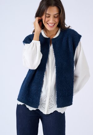 Lachende vrouw met een navyblauw, gestructureerd mouwloos vest over een witte blouse met knopen en geperforeerde zoom, gecombineerd met donkerblauwe jeans.