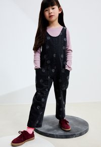 Salopette en denim noir à pois gris, avec poches devant, portée sur un haut à manches longues rose clair. Associée à des chaussures en daim bordeaux et des chaussettes roses.