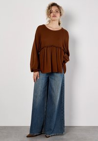 Camisa marrón de manga larga con canalé y cintura con volantes, combinada con unos vaqueros de denim azules desgastados y de pierna ancha. Larga hasta el suelo y de corte relajado.