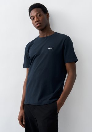 Νεαρός άνδρας με σκούρο δέρμα, που φοράει μπλε ναυτικό μπλουζάκι BOSS και μαύρο παντελόνι, στέκεται μπροστά σε λευκό τοίχο με το ένα χέρι στην τσέπη.