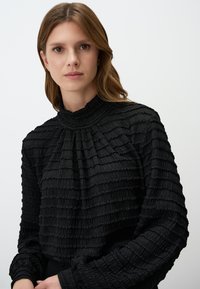 Zwarte blouse met textuur, een hoge, geraffineerde kraag en lange mouwen. Heeft horizontale plooien en een ontspannen pasvorm.