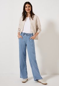Jasno beżowa lniana koszula o luźnym kroju, zestawiona z luźnymi jasnoniebieskimi dżinsami i białymi trampkami. Spodnie mają elastyczny pas.