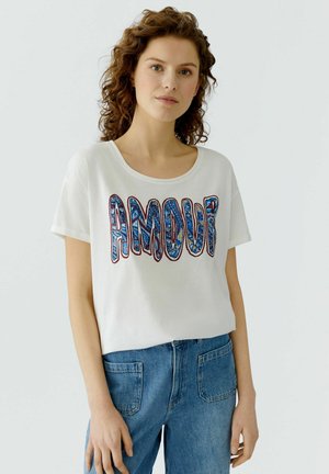 Junge Frau mit lockigem Haar, die ein weißes T-Shirt mit dem Schriftzug "AMOUR" in blauen Mustern trägt, kombiniert mit hoch taillierten blauen Denim-Jeans.