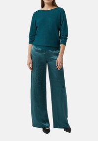 Kobieta w turkusowej długiej bluzie dzianinowej i błyszczących szerokich turkusowych spodniach, stojąca z rękami swobodnie przy boku.