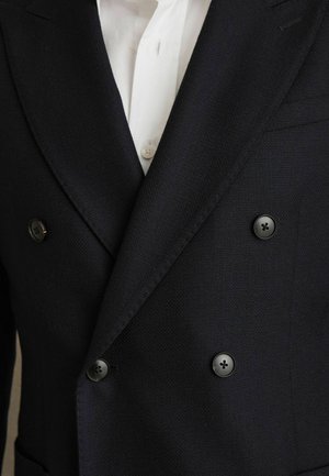 Blazer bleumarin cu două rânduri de nasturi, dintr-un material texturat, cu accente de nasturi negri, purtat alături de o cămașă albă parțial vizibilă dedesubt.