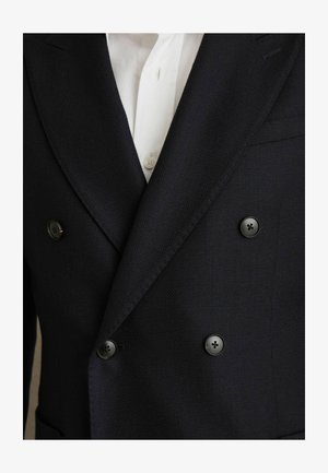 Dvojité sako v námořnické barvě z texturované tkaniny s černými knoflíky, v kombinaci s bílou košilí, která je částečně viditelná pod sako.