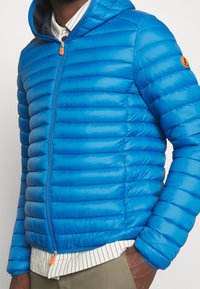 Giacca a vento blu con baffle orizzontali, chiusura con zip e cappuccio. Presenta un tirazip arancione e un emblema del marchio sulla manica.
