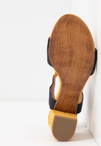 Keltas ja musta sandaali, millel on sulepehme ülemine osa. Omab paksu puidust kantimist, tekstuurset kummist talda ja ümarat varba disaini.