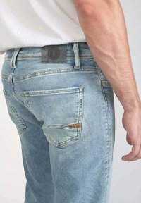 Los jeans de mezclilla azul claro presentan un bolsillo trasero con un acento marrón y detalles de costura. La cinturilla tiene una etiqueta oscura y costuras visibles.