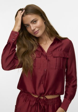 Burgundy satin blouse with a button-up front, two chest pockets, long sleeves, and a cropped design featuring a tied waist. Smooth texture.