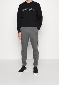 Svart sweatshirt med vit grafisk text, parat med grå träningsbyxor med sidostråk och en logotyp. Bärs med svarta sneakers.