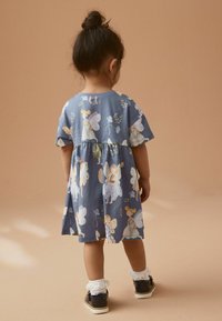 Kurzes, blaues Baumwollkleid mit floralem und Feen-Print, gerafftem Taillenbund und knielangem Saum. Kombiniert mit dunklen Schuhen und Socken.