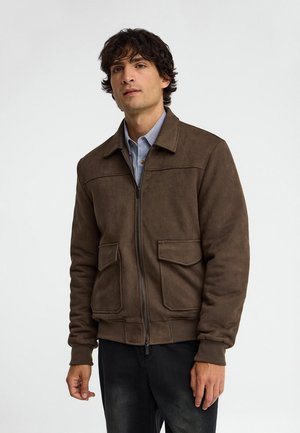 Uomo che indossa una giacca marrone con zip e tasche con patta sopra una camicia blu con colletto, in piedi davanti a uno sfondo chiaro uniforme.