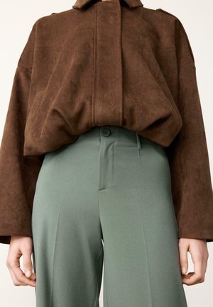 Kestenjak od smeđeg antilopa s velikim ovratnikom uparen s visokim zelenim hlačama s jednom kopčom i urednim prednjim naborkama.