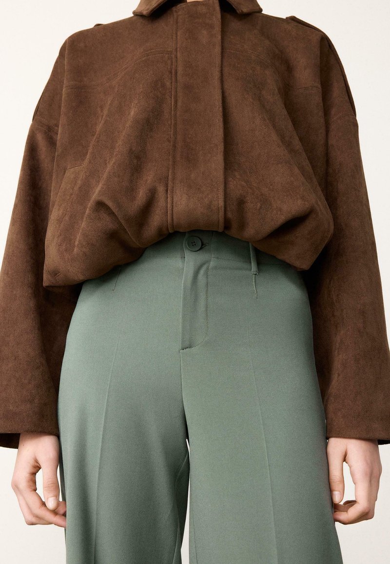 Veste oversize en daim marron avec col, associée à un pantalon taille haute vert, doté d'un bouton unique et de plis avant nets.