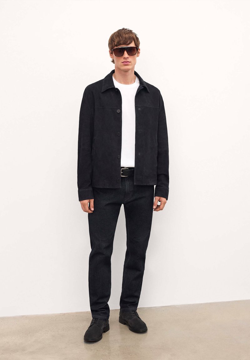 Chaqueta de ante negra sobre una camiseta blanca, combinada con pantalones oscuros y zapatos negros. Grandes gafas de sol rectangulares completan el look.