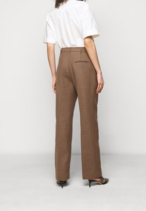 Persona con pantalones marrones de cuadros de tiro alto y pierna ancha, camisa blanca de manga corta y zapatos negros de tacón con punta afilada, de pie de espaldas.