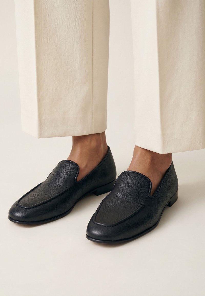 Sorte læder slip-on loafers med off-white vidde bukser, der viser ankler mod en ensfarvet lys baggrund.