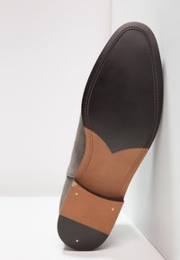 Semelle de chaussure en daim gris avec une section contrastante en cuir fauve. Comprend une semelle extérieure en caoutchouc noir et des coutures décoratives le long du bord.