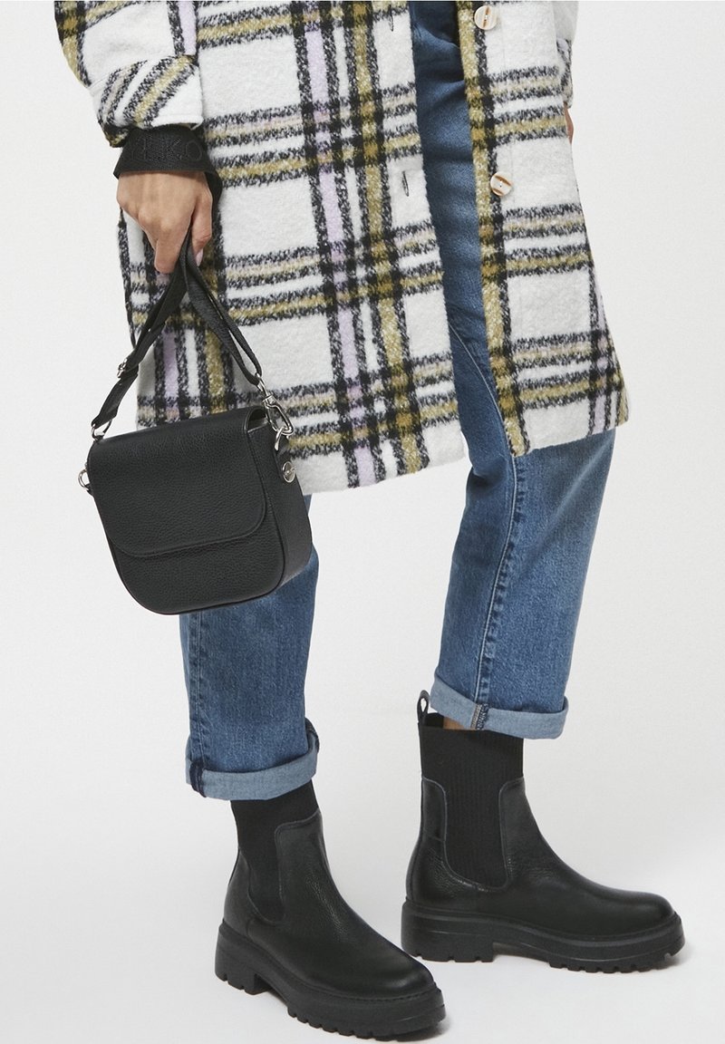 Personne portant un jean bleu retroussé, des bottines noires épaisses, tenant un petit sac à main noir et portant un manteau à carreaux blanc avec des lignes jaunes et noires.