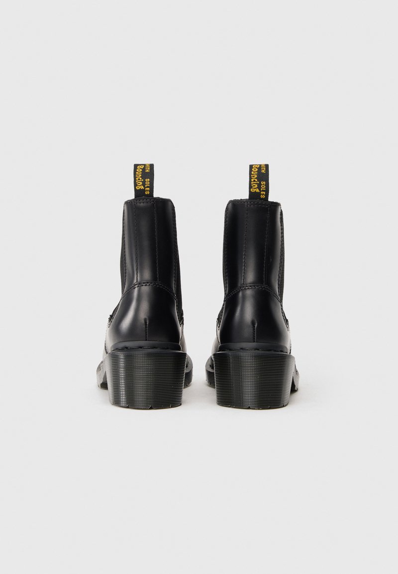 Un par de botas de cuero negro hasta el tobillo con tacones estriados y tiradores, con texto amarillo, vistas desde la parte trasera sobre un fondo liso.