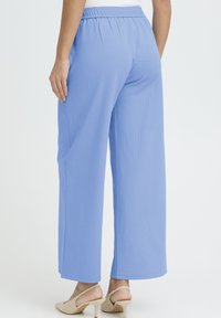 Svetlo modre hlače širokih hlačnic z elastičnim pasom. Gladek, lahek material. Zaključeni robovi in stranski žepi. Nošene z bež petami.