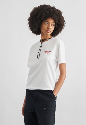 Donna con capelli afro che indossa una t-shirt bianca Carhartt WIP e pantaloni neri, in piedi con una mano in tasca davanti a uno sfondo neutro.