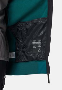 Interno di giacca nera con rivestimento in pile teal. Include etichette in tessuto resistente all'acqua e traspirante e un orlo testurizzato con zip.