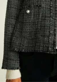 Schwarze und weiße Tweedjacke mit fransigen Kanten, ausgestattet mit einem runden Perlenknopf. Der Ärmel zeigt ein weißes Strickbündchen darunter.