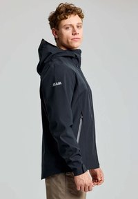 Giacca impermeabile nera con cappuccio, dotata di tasche con zip e design aderente. Accenti in grigio sui pannelli laterali. Logo sulla manica.