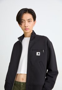 Giacca nera con zip realizzata in tessuto testurizzato, con una tasca sul petto dotata di zip e una toppa con logo. Indossata sopra una camicia bianca.