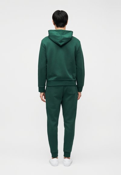 Άνδρας με σκουροπράσινο φούτερ με κουκούλα και αντίστοιχο παντελόνι φόρμας, στέκεται με την πλάτη γυρισμένη, φοράει άσπρα αθλητικά παπούτσια σε απλό ανοιχτό φόντο.