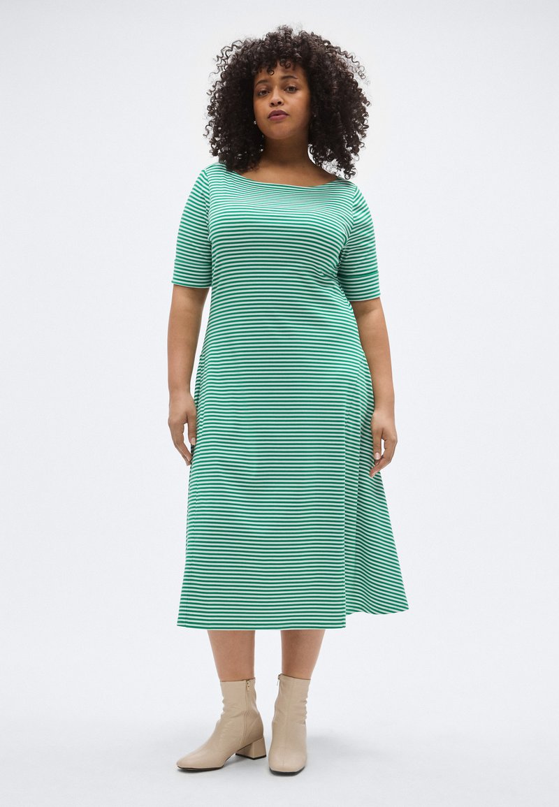 Zöld-fehér csíkos ruha rövid ujjakkal és széles nyakkivágással, áramló kialakítással és texturált anyagból. Bézs bokacsizmával viselve.