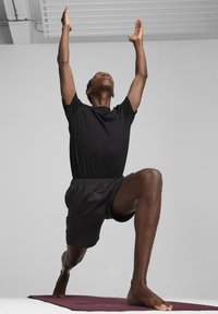 Sort atletisk t-shirt og shorts iført af en person i lungeposition på et bordeaux yoga tæppe, med armene strakt opad.