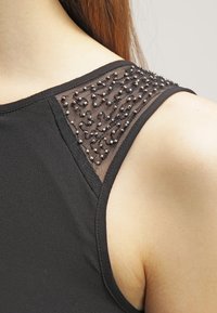 Černý top bez rukávů s průhledným detailem ramene zdobeným malými kulatými ozdobami, které dodávají texturu a kontrast.