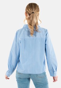 Camicia button-up azzurra chiara con sottili righe bianche, maniche lunghe e vestibilità rilassata. Presenta bottoni sui polsini e un dettaglio sulla schiena.