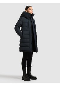 Mornarsko plava naduvana jakna s visokim, prostranim kapuljačom, prošivenim dizajnom, gumbima sprijeda i bočnim džepovima, uparena s crnim čizmama do koljena i tajicama.