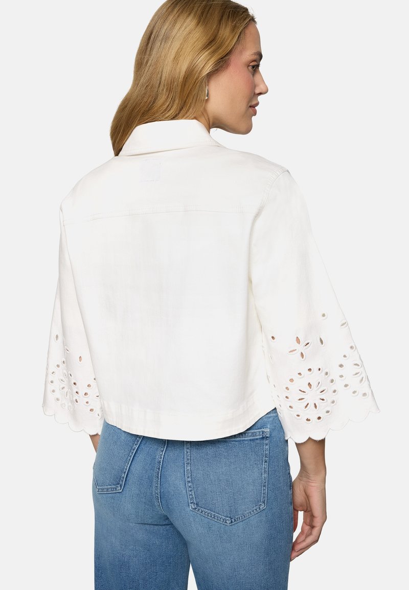 Femme aux cheveux blonds portant un chemisier blanc avec des découpes florales ajourées sur des manches larges aux trois quarts et un jean bleu, vue de dos sur fond blanc.