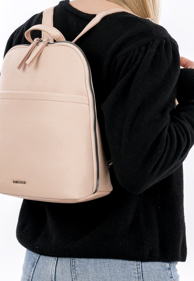 Sac à dos en cuir beige avec fermeture éclair porté à l'épaule par une personne vêtue d'un pull noir et d'un jean bleu clair.