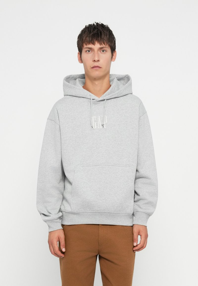 Sudadera gris con un bolsillo frontal, capucha con cordón y logo de GAP en blanco. Combinada con pantalones marrones, con puños y dobladillo acanalados.