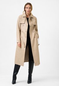 Beige leather trench coat with double-breasted design, large collar, and decorative buttons. Paired with black fitted clothing and knee-high boots.