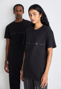Camisetas de algodón negras con corte relajado, que presentan un estampado de logo en blanco. También son visibles unos pantalones grises texturizados que lleva puestos el modelo en frente.