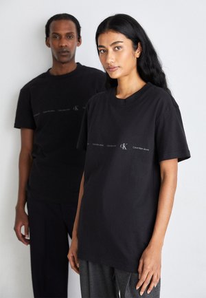 Camisetas de algodón negras con corte relajado, que presentan un estampado de logo en blanco. También son visibles unos pantalones grises texturizados que lleva puestos el modelo en frente.