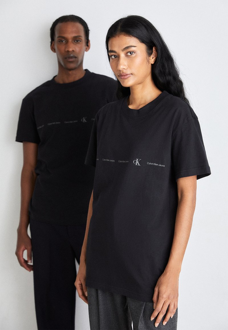 Camisetas de algodón negras con corte relajado, que presentan un estampado de logo en blanco. También son visibles unos pantalones grises texturizados que lleva puestos el modelo en frente.
