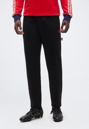 Bărbat purtând pantalonii de trening negri Adidas cu logo roșu, bluză cu mâneci lungi roșu-albastru și ghete negre de fotbal cu accente verde neon.