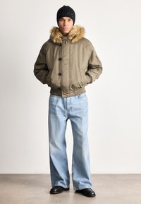 Hombre vestido con una chaqueta de khaki con capucha de pelo, un gorro de punto negro con logo, jeans de pierna ancha de color azul claro y zapatos negros, de pie contra un fondo liso.