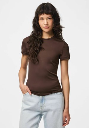 Mujer con el pelo largo, oscuro y rizado, que lleva una camiseta marrón ajustada de manga corta y vaqueros azul claro, de pie con una mano en el bolsillo.